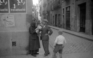 Fotografia antiga d'un carrer de Gràcia amb dona venent diaris que xerra amb un home i un nen