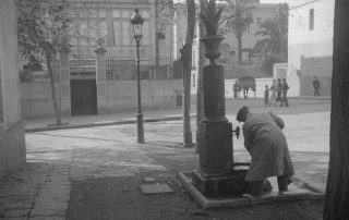 Fotografia antiga en blanc i negre d'un home al costat d'una font d'un carrer de Gràcia