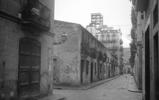 Fotografia antiga en blanc i negre d'un carrer amb un vianant i un gat negre