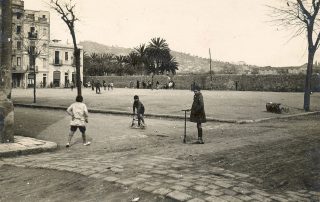 Fotografia antiga en blanc i negre d'uns nens jugant a una plaça amb patinet i bicicleta