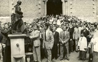 Fotografia antiga blanc i negre amb gent davant d'una monument al costat d'una esglèsia de Gràcia