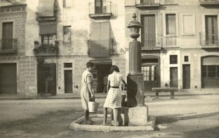 Fotografia antiga en color sèpia d'una dona i una noia prenent aigua d'una font a una plaça